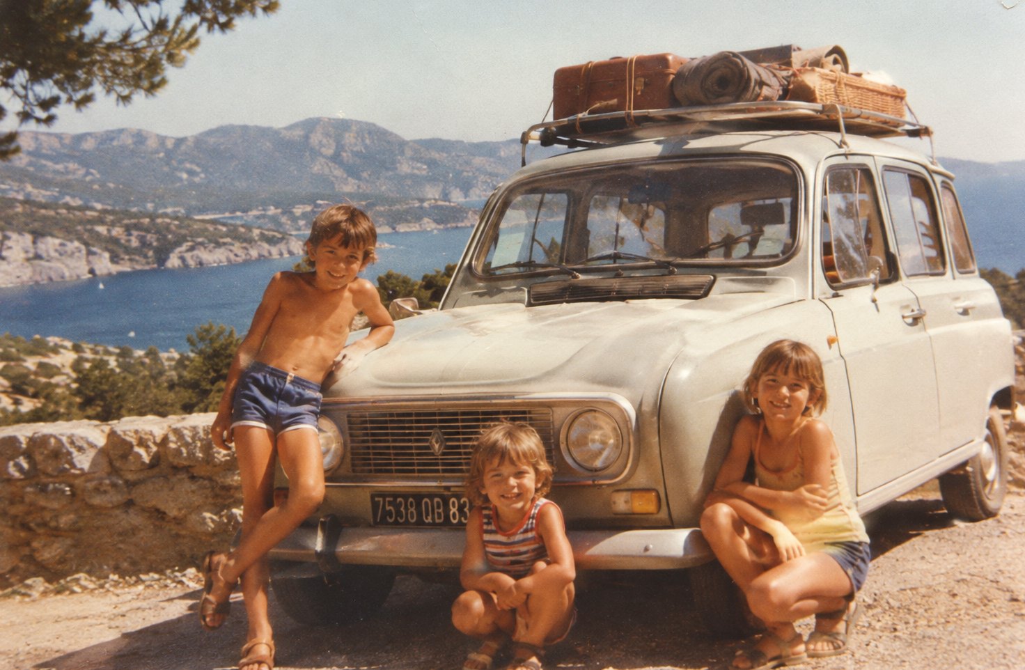 Une photo d'époque : trois enfants posent devant une 4L chargée de bagages, au-dessus de la mer
